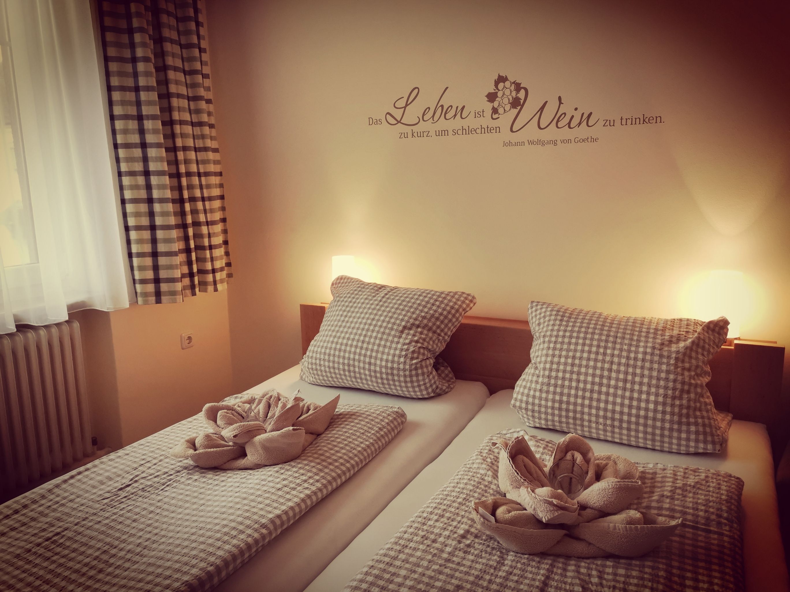 Hangulatos vendégszoba két ággyal, kockás paplanokkal és párnákkal, dekoratív törölközőkkel és egy Goethe-idézettel a falon.
