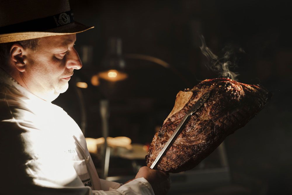 Egy fehér inges férfi egy nagy, grillezett húsdarabot tart késsel a kezében.