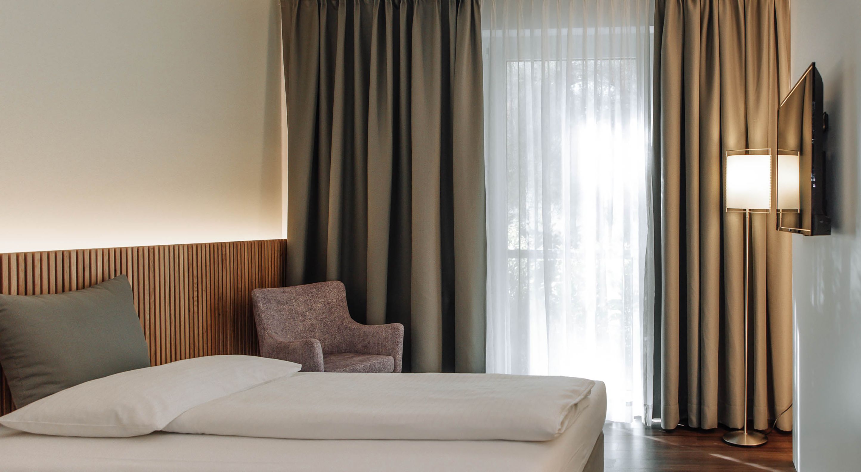 Modern szállodai szoba ággyal, karosszékkel és függönyökkel.