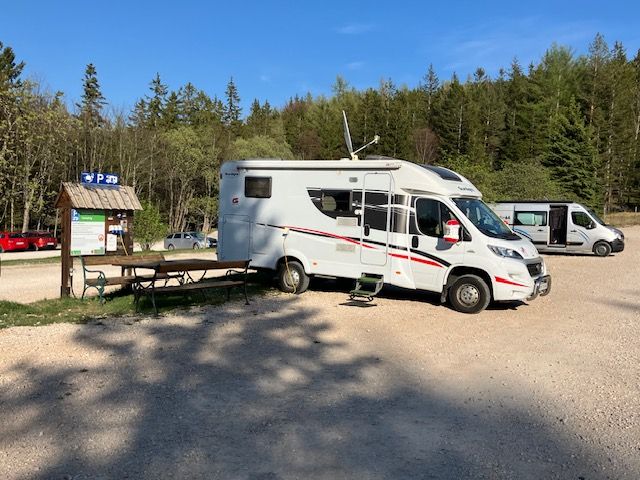 Lakóautó a Hohe Wand natúrpark parkolójában, fákkal és kék éggel körülvéve.