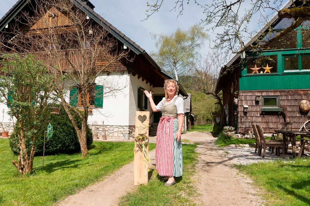 Egy hagyományos ruhás nő áll két rusztikus épület előtt egy zöld udvaron.