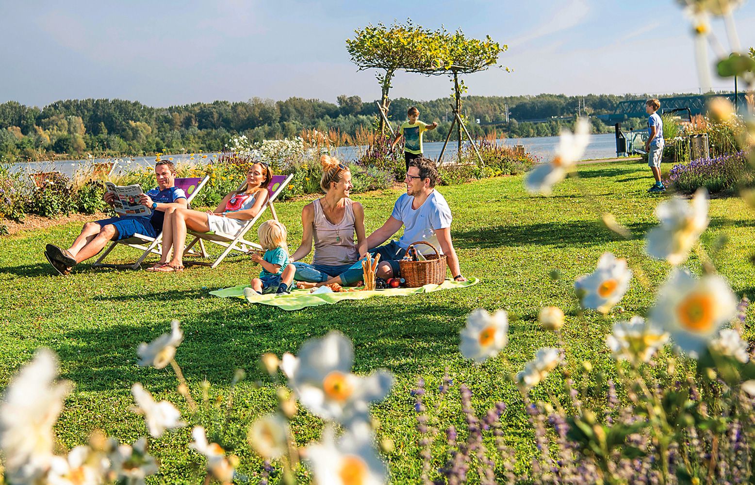 Családi piknik vidéken, virágokkal az előtérben és egy folyóval a háttérben.