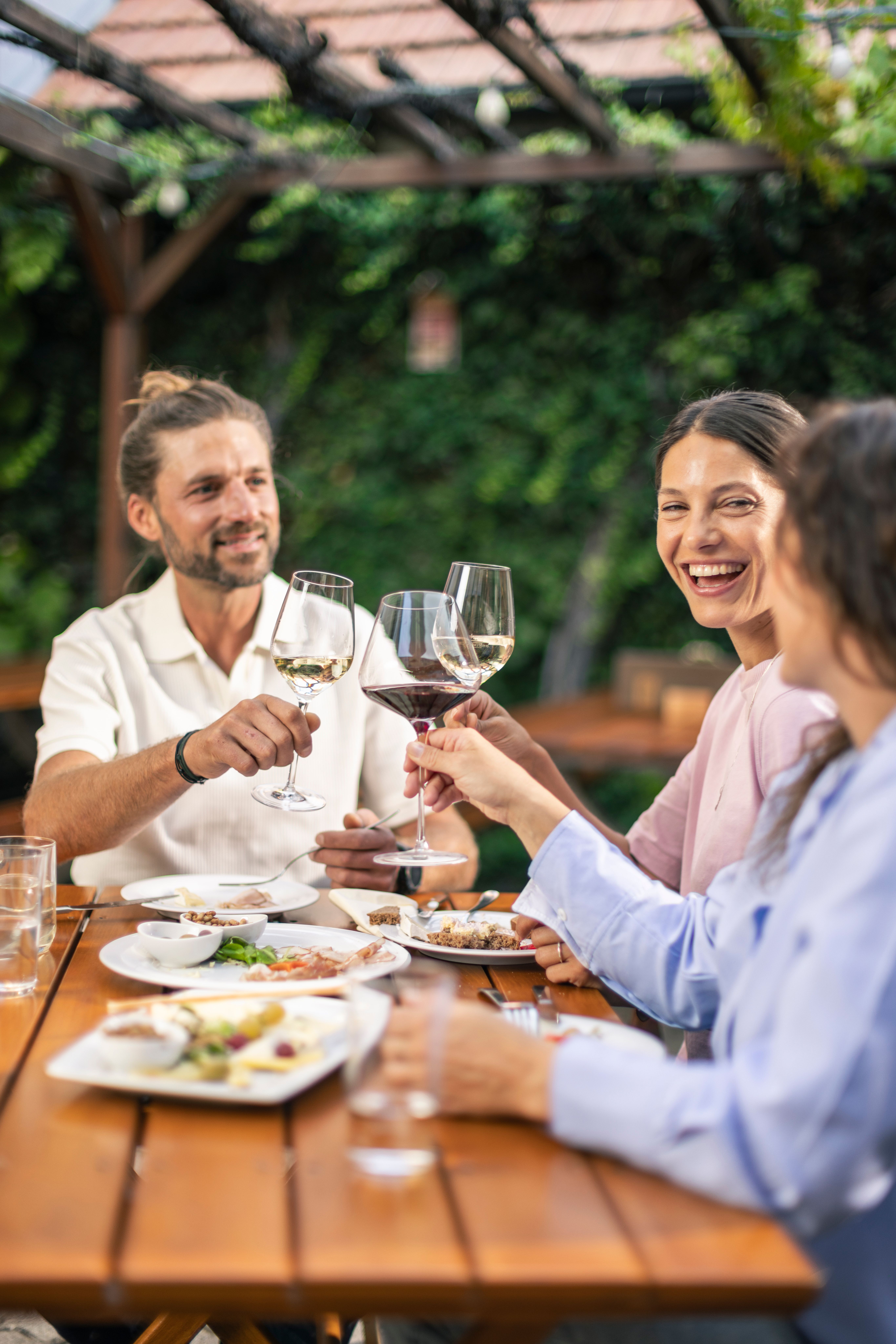 Drei Gäste stoßen im Gastgarten des Weinguts Herzog in Bad Vöslau mit Wein an – niederösterreichische Heurigenkultur mit Jause und entspannter Atmosphäre im Wienerwald.