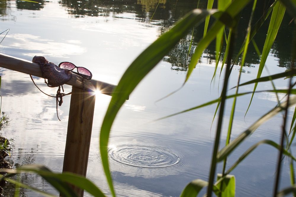 Egy fakorlát egy tóparton, rajta napszemüveg és bikini. A vízben kis hullámok láthatók, nádasokkal körülvéve.