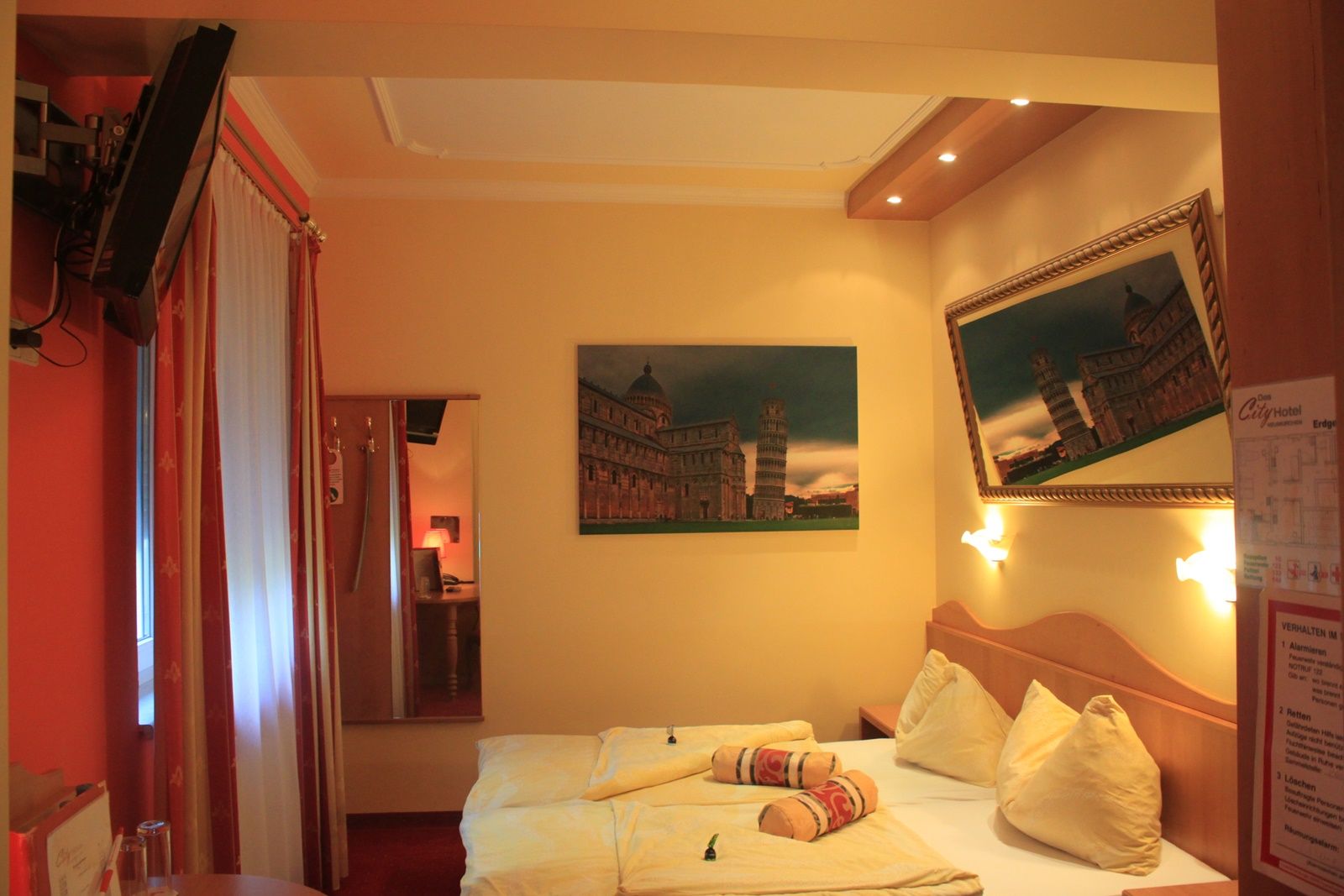 Hotelszoba franciaággyal, a pisai ferde torony képeivel és egy TV-vel a falon.