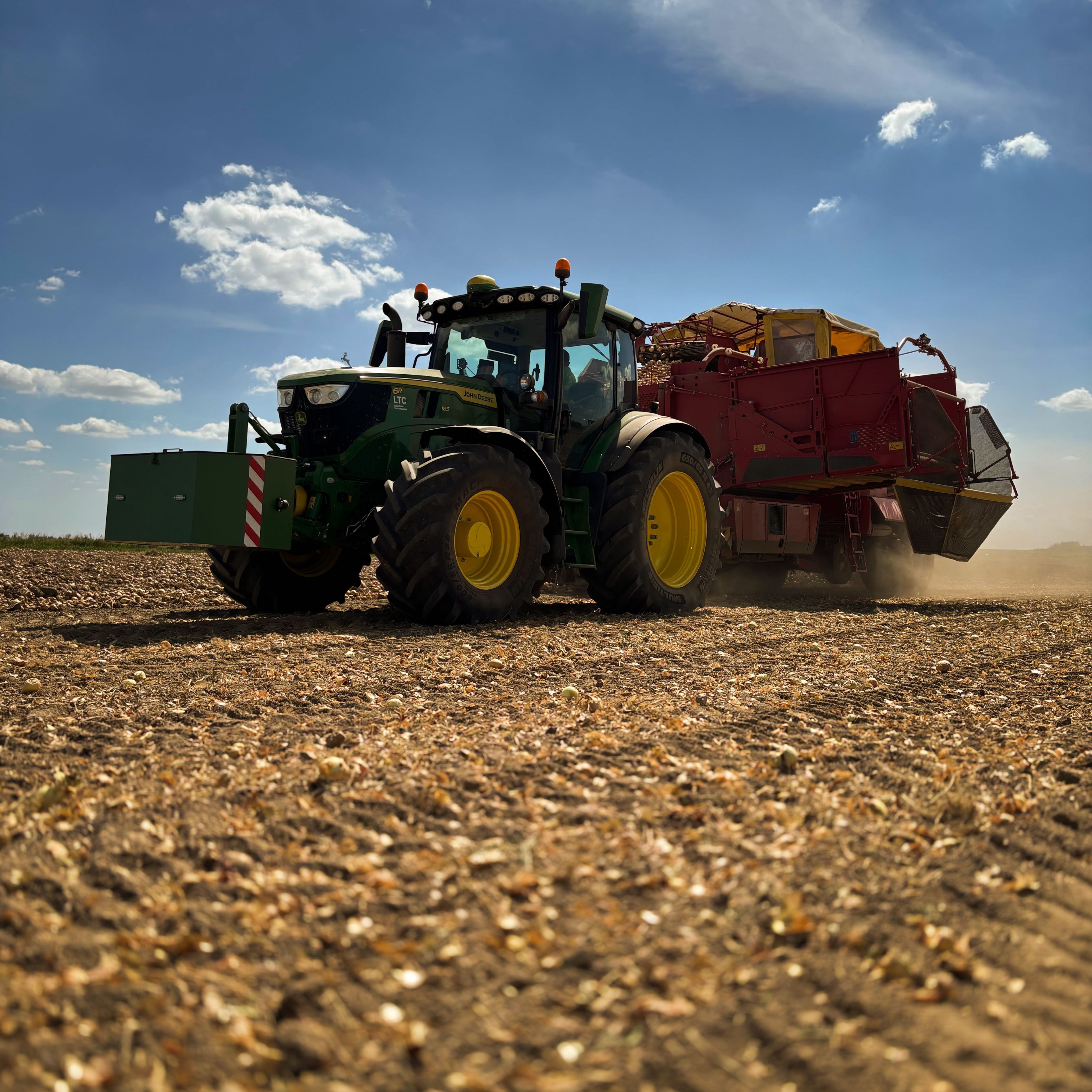 Egy traktor pótkocsival halad át egy betakarított mezőn, kék égbolt alatt, néhány felhővel.