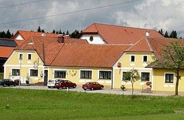 Sárga épület piros tetővel és több autóval előtte, zöld rétekkel és fákkal körülvéve.