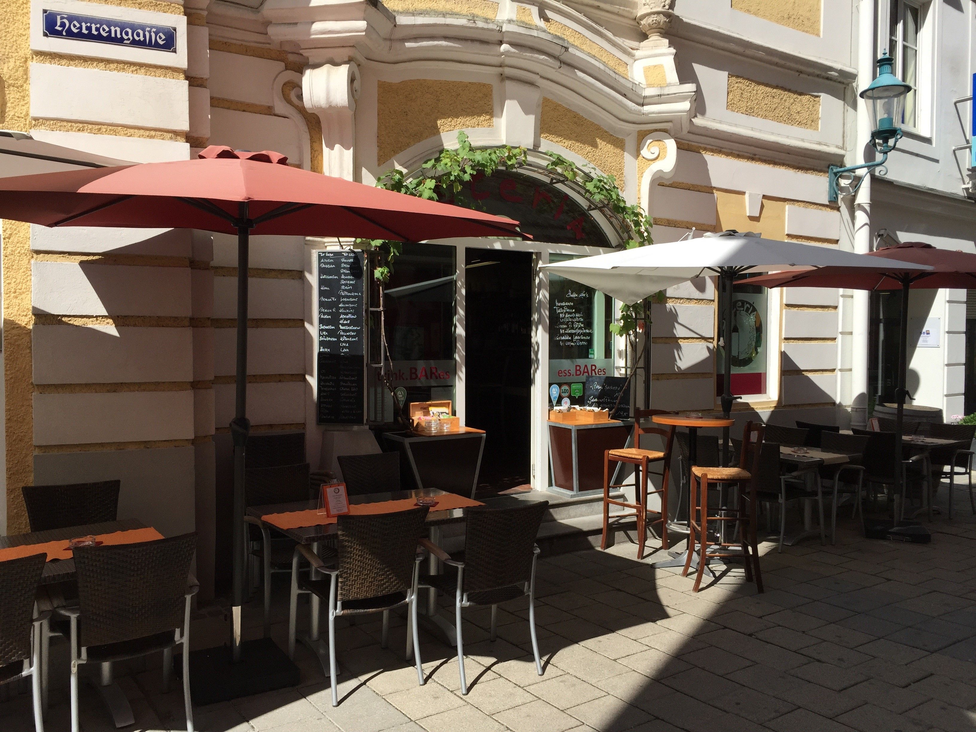 Egy étterem szabadtéri területe asztalokkal, székekkel és napernyőkkel egy napsütötte sikátorban.