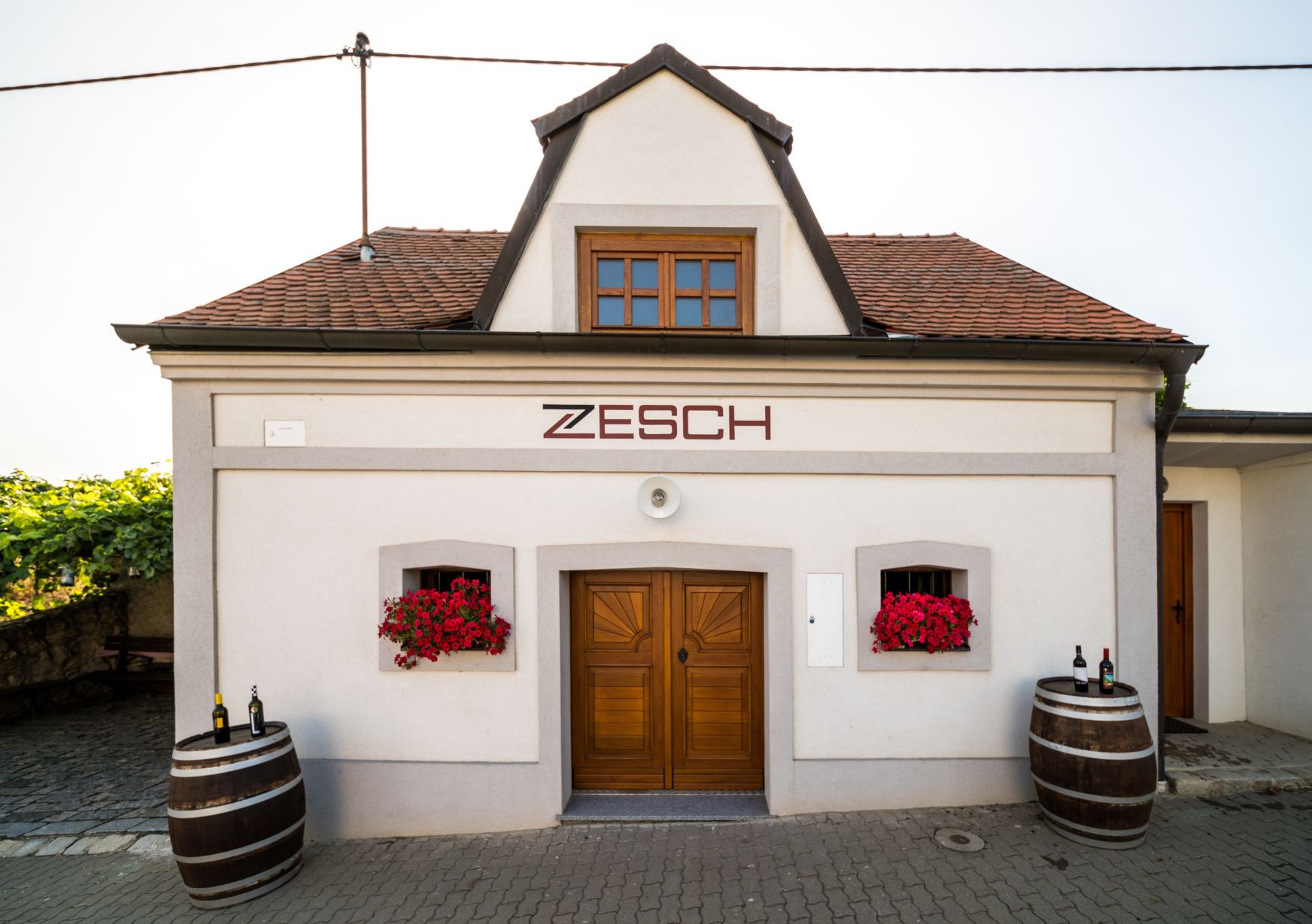 Kis épület "Zesch" felirattal, két boroshordóval és piros virágokkal.