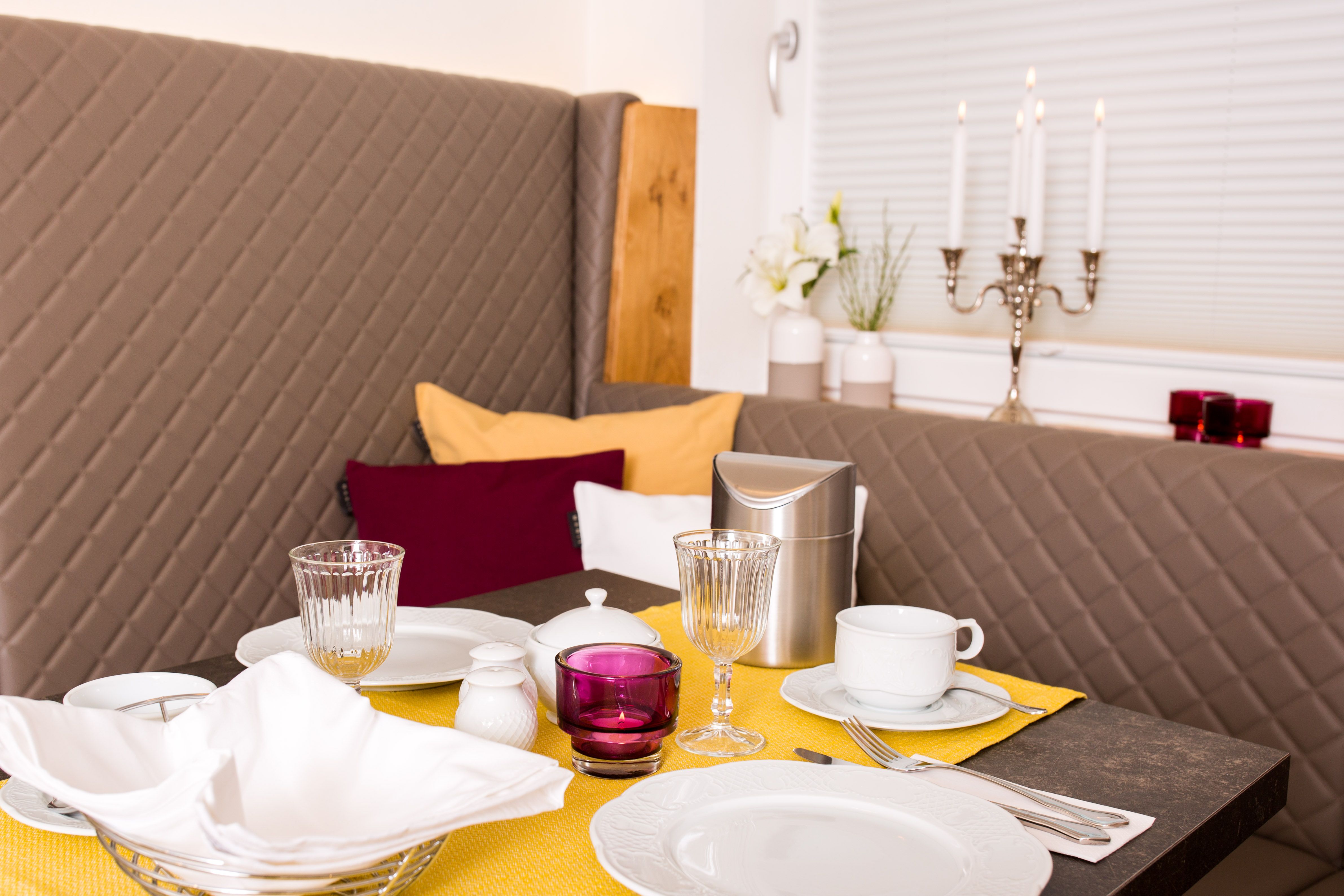 Elegáns reggelizőasztal sárga futóval, edényekkel, poharakkal és gyertyatartókkal.