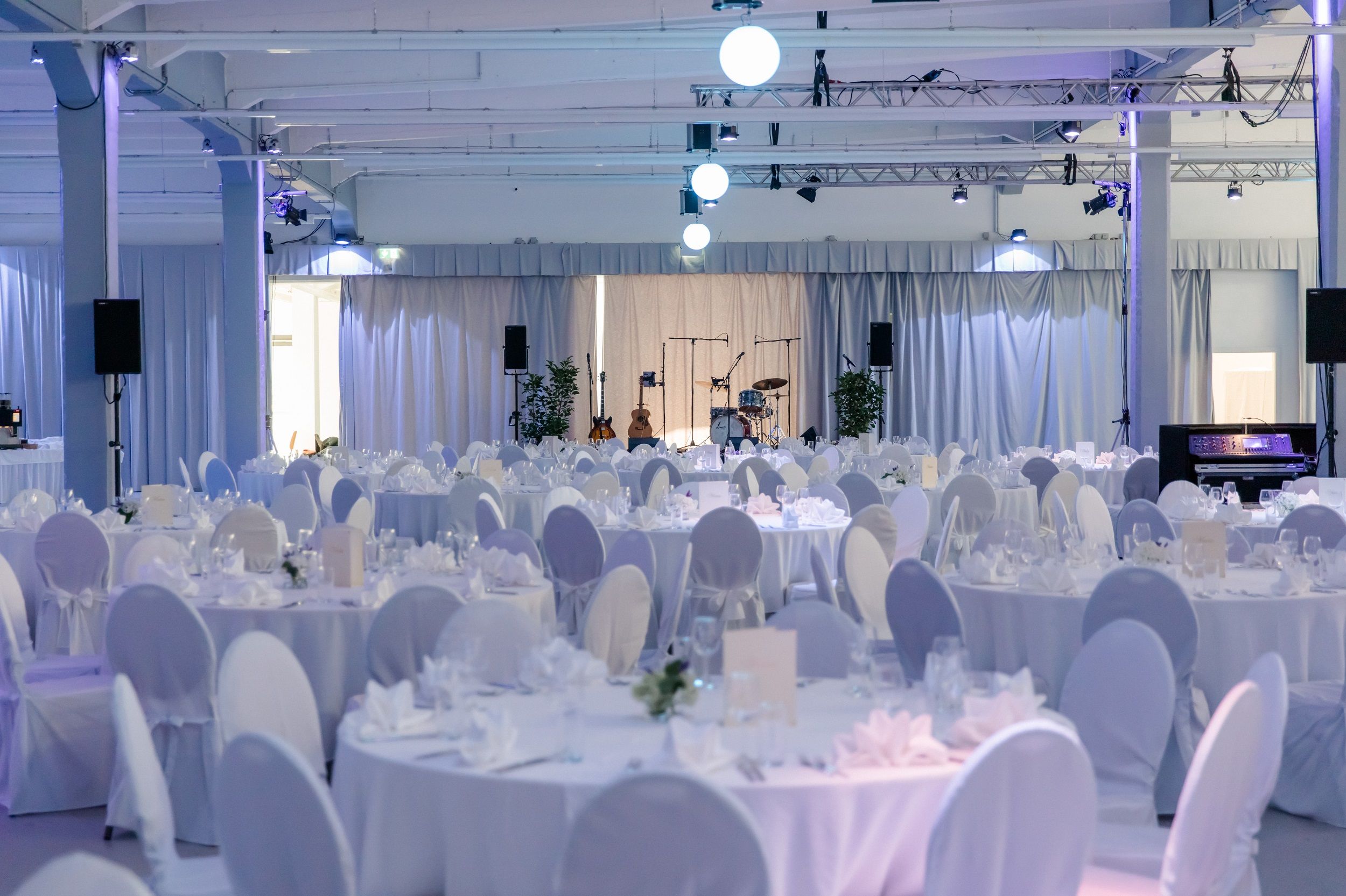Elegáns rendezvényterem kerek, fehér abroszos asztalokkal és hangszerekkel felszerelt színpaddal.