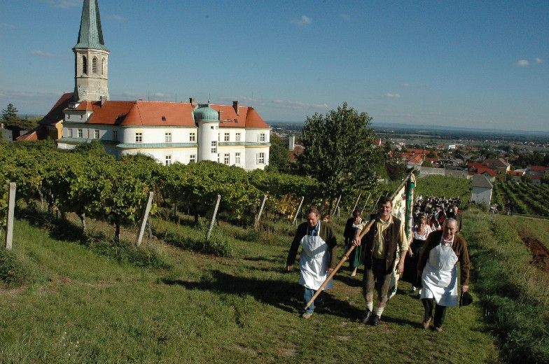 Hagyományos ruhába öltözött emberek sétálnak a gumpoldskircheni szőlőskertekben, a háttérben egy templom és épületek.