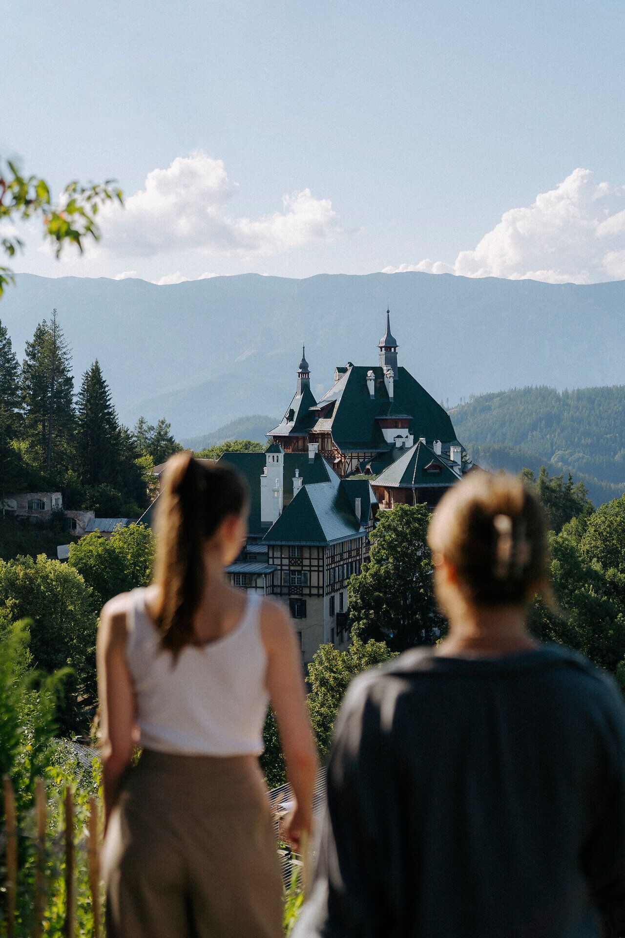 Két ember, háttal a kamerának, egy festői hegyvidéki tájra néz. Az előtérben a Südbahnhotel látható, buja zöldövezetben. A háttérben hegyek emelkednek a tiszta kék ég alatt.