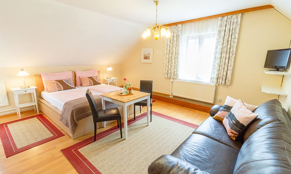 Hangulatos vendégszoba franciaággyal, kanapéval, asztallal és székekkel, világos berendezéssel.