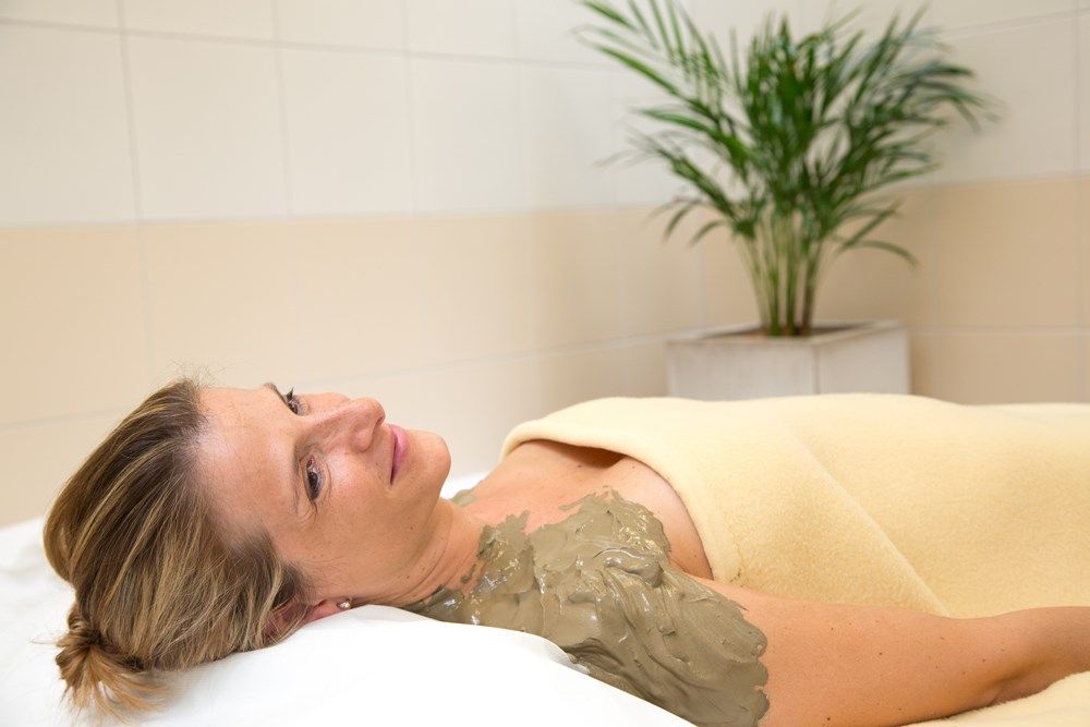 Nő pihen kénes iszappakolással a vállán, takaróba burkolózva, a wellness-részlegben, háttérben egy növény.