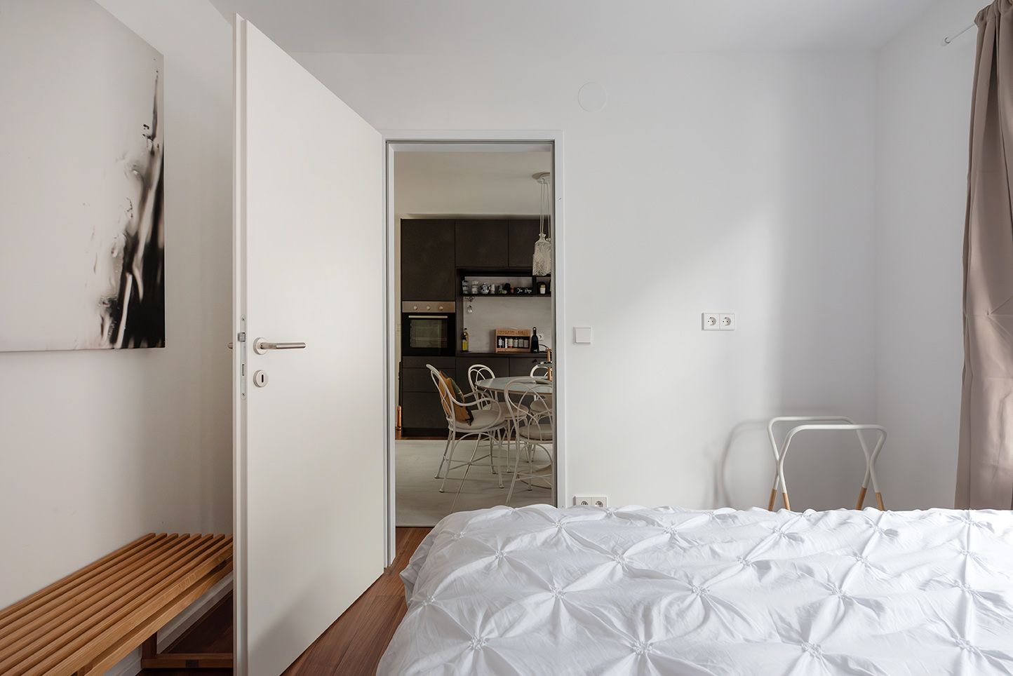 Modern hálószoba fehér ágyneművel, nyitott ajtóval, amely az étkezőbe vezet, és minimalista bútorokkal.
