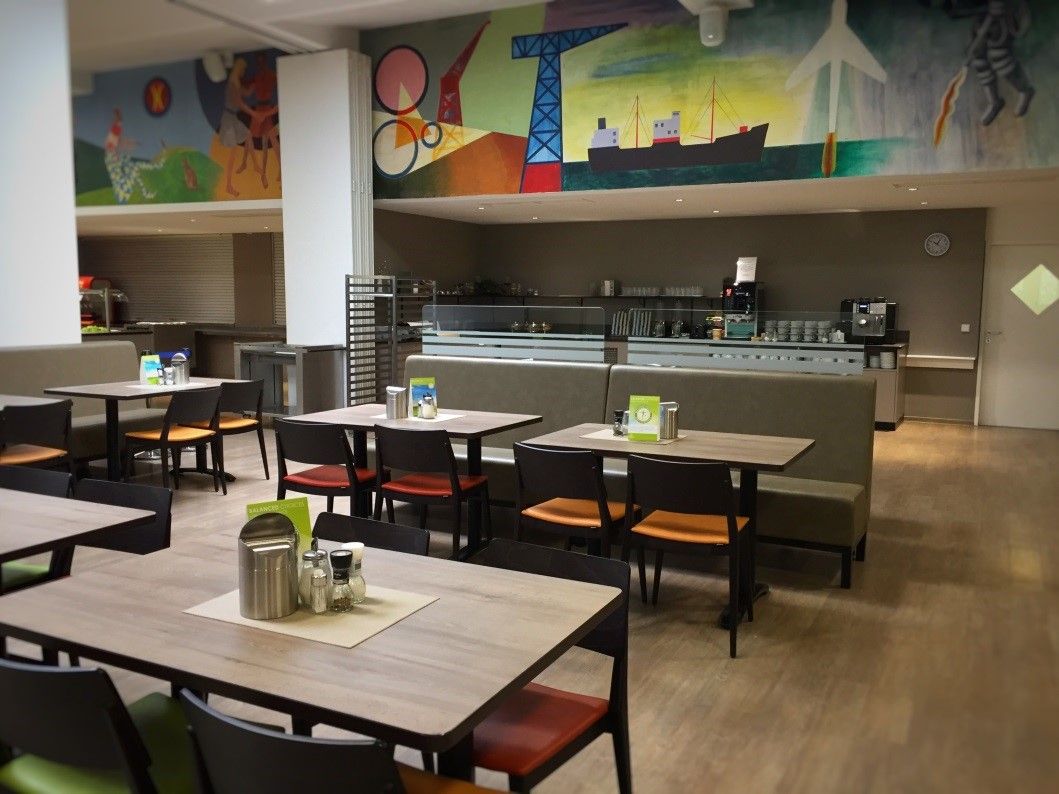 Egy étterem belső nézete üres asztalokkal és székekkel, a háttérben színes falfestmény.