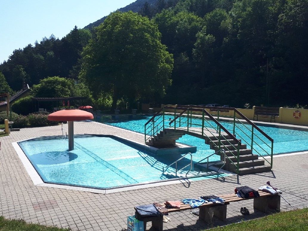 Kültéri medence egy kis medencével, egy híddal és egy gombás vízjátékkal. Fákkal és napsütötte égboltal körülvéve.