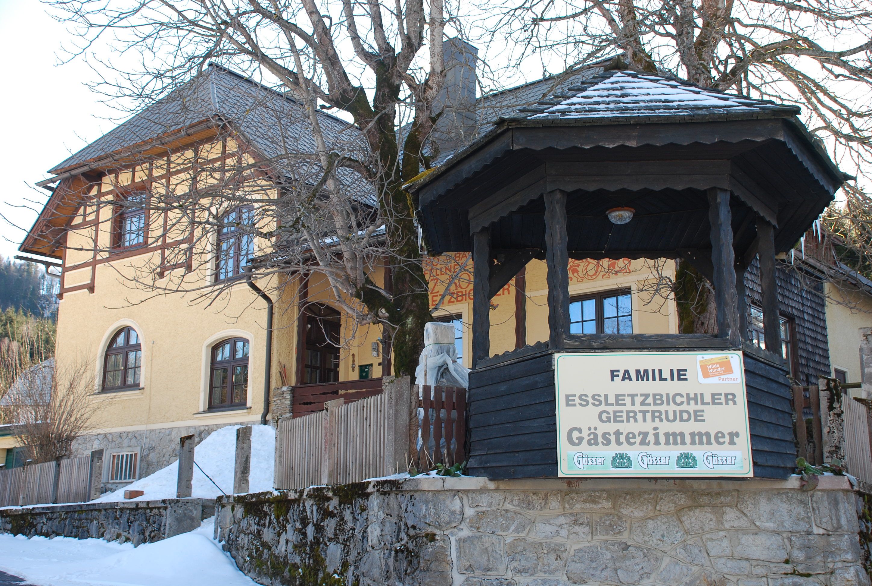Sárga vendégház fából készült verandával és "Familie Essletzbichler Gertrude Gästezimmer" felirattal.
