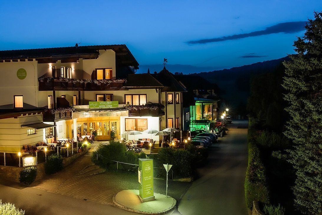 A Vitalzeit Hotel Weber külső nézete éjszaka, meleg fénnyel megvilágítva, fákkal és parkoló autókkal körülvéve.
