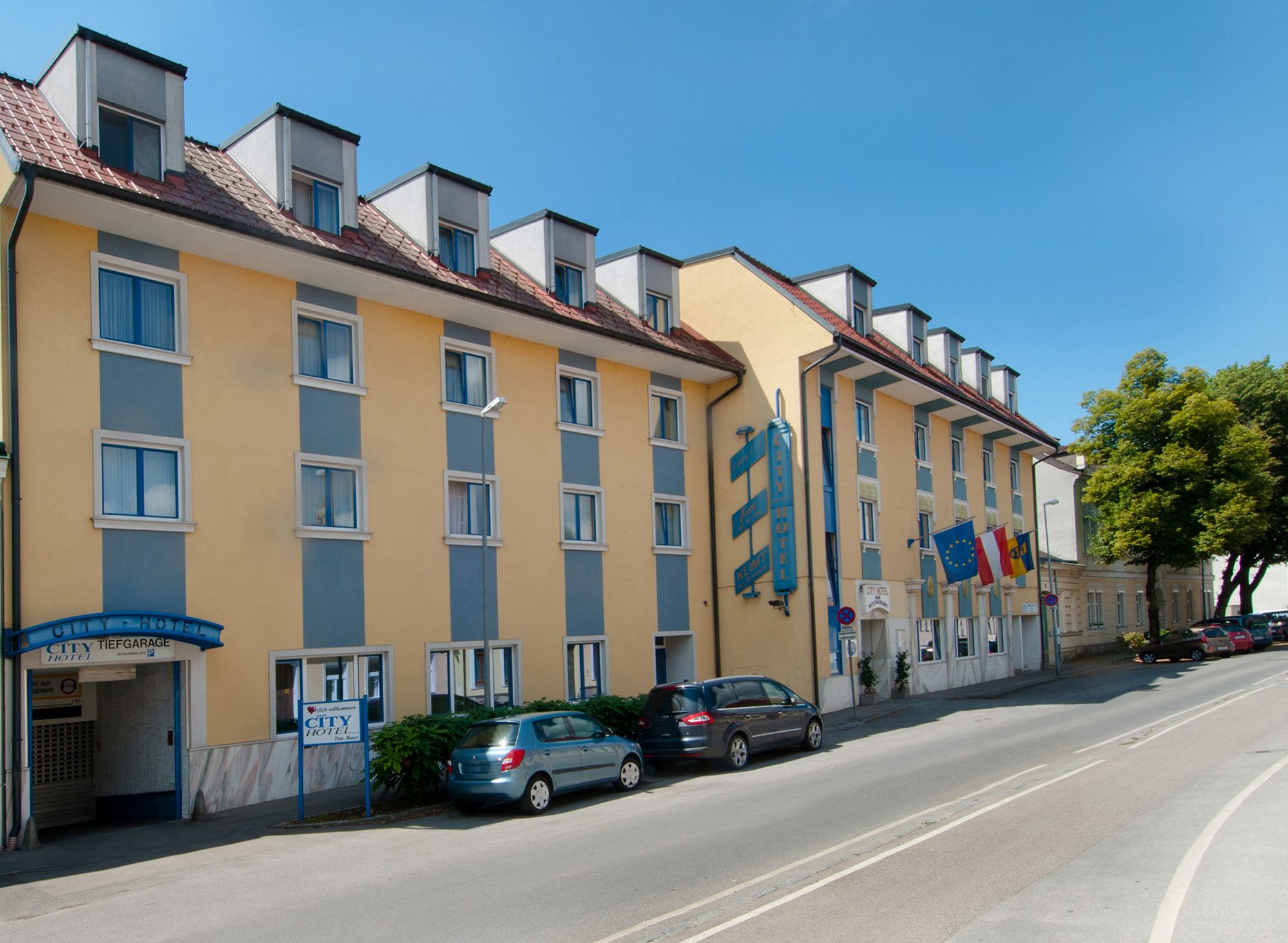 Egy sárga és kék színű szálloda külső nézete, több ablakkal és parkoló autókkal előtte.
