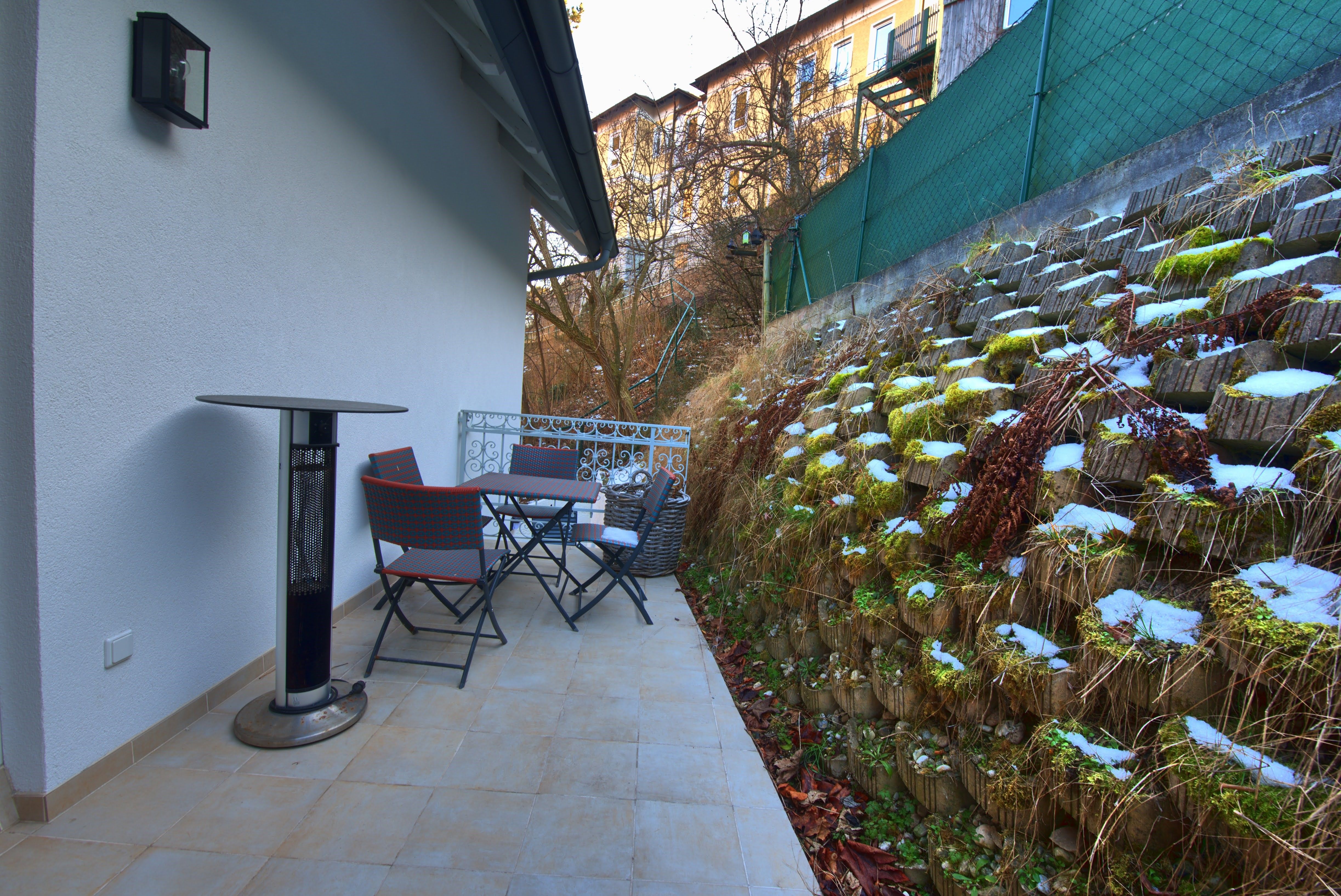 Terasz asztallal, székekkel és hősugárzóval egy zöld fal mellett.