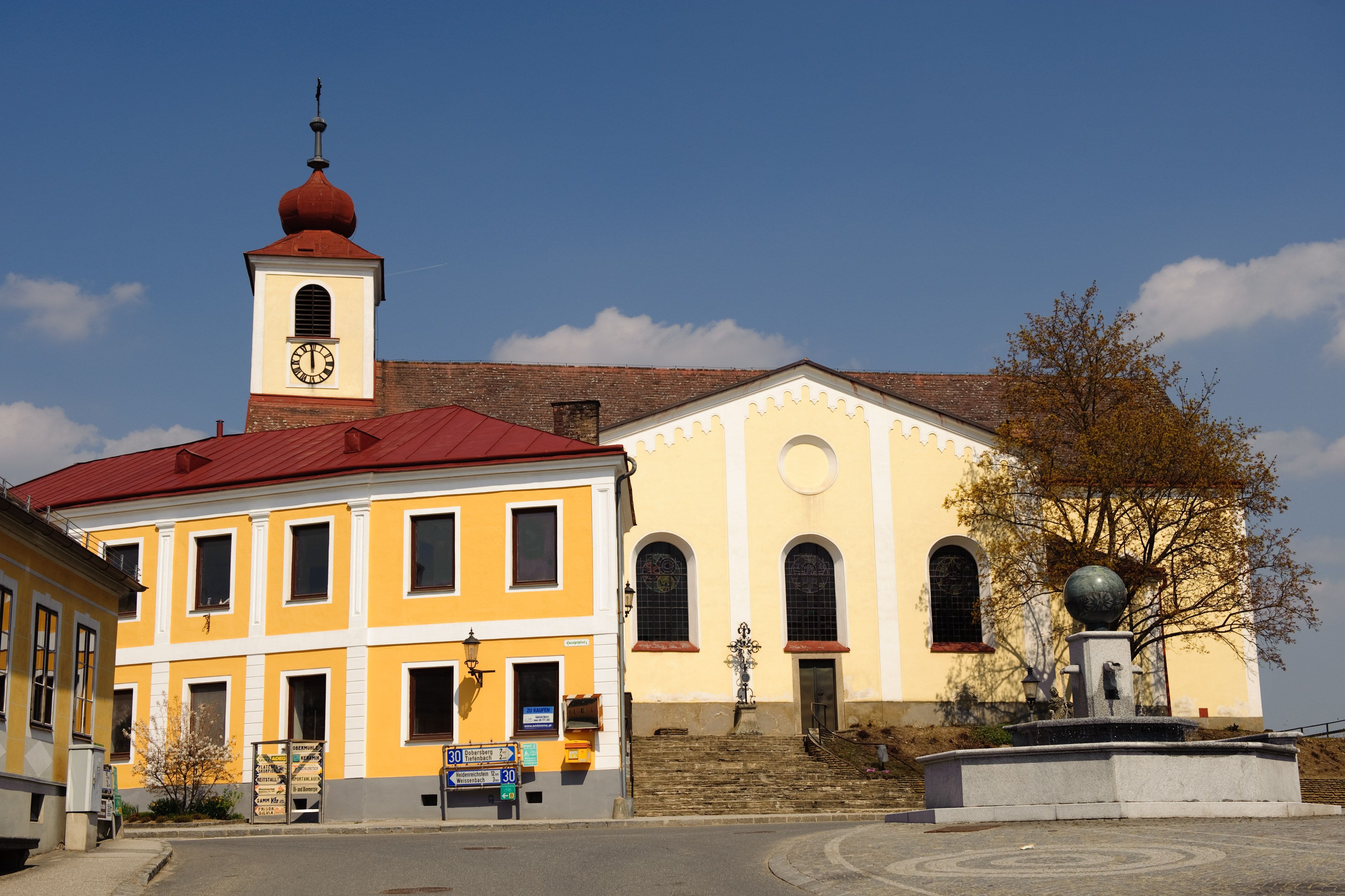 Sárga homlokzatú templom, piros tetejű toronnyal a főtéren.