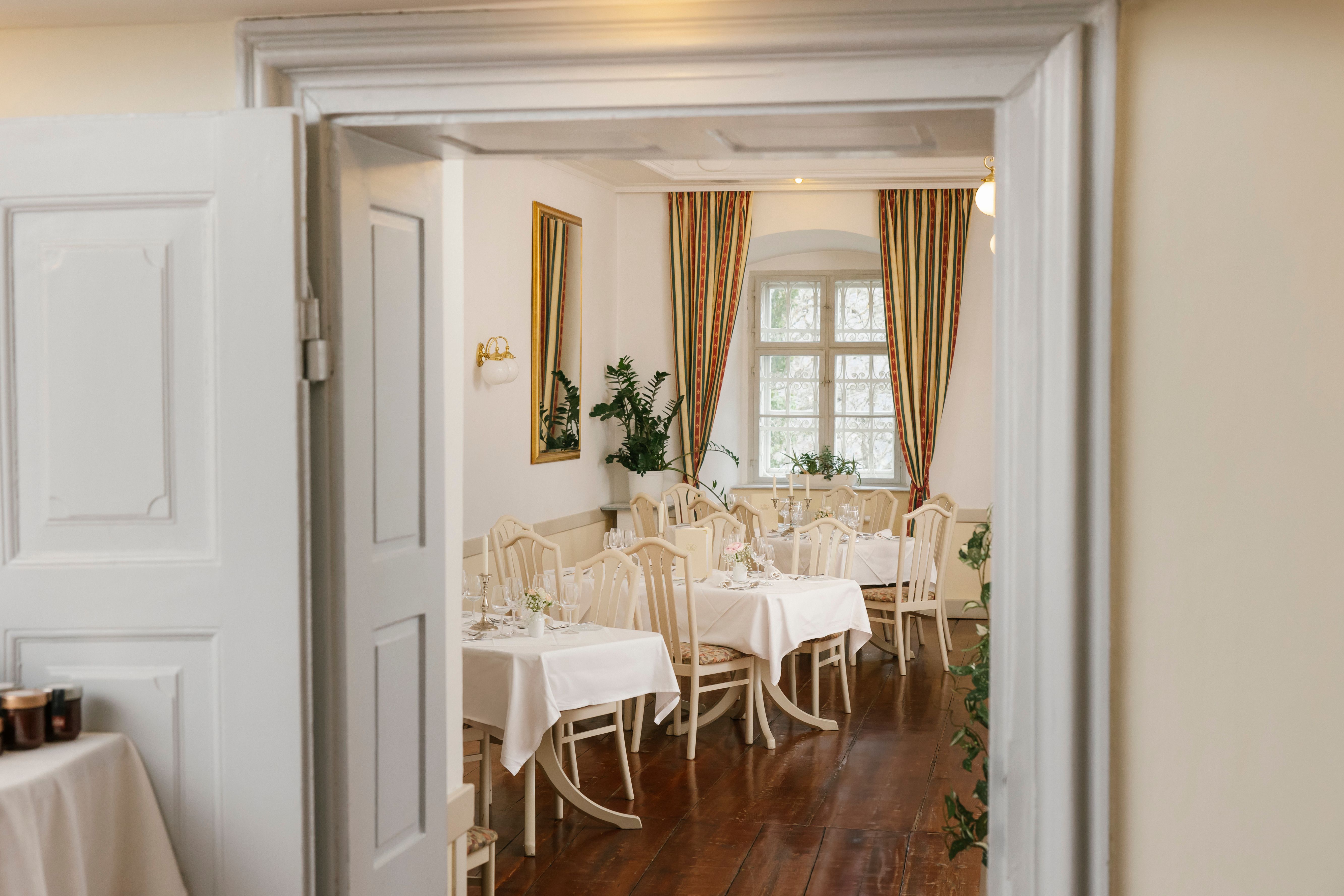 Elegáns étterem terített asztalokkal, fehér terítőkkel és klasszikus székekkel, nyitott ajtón keresztül látható.