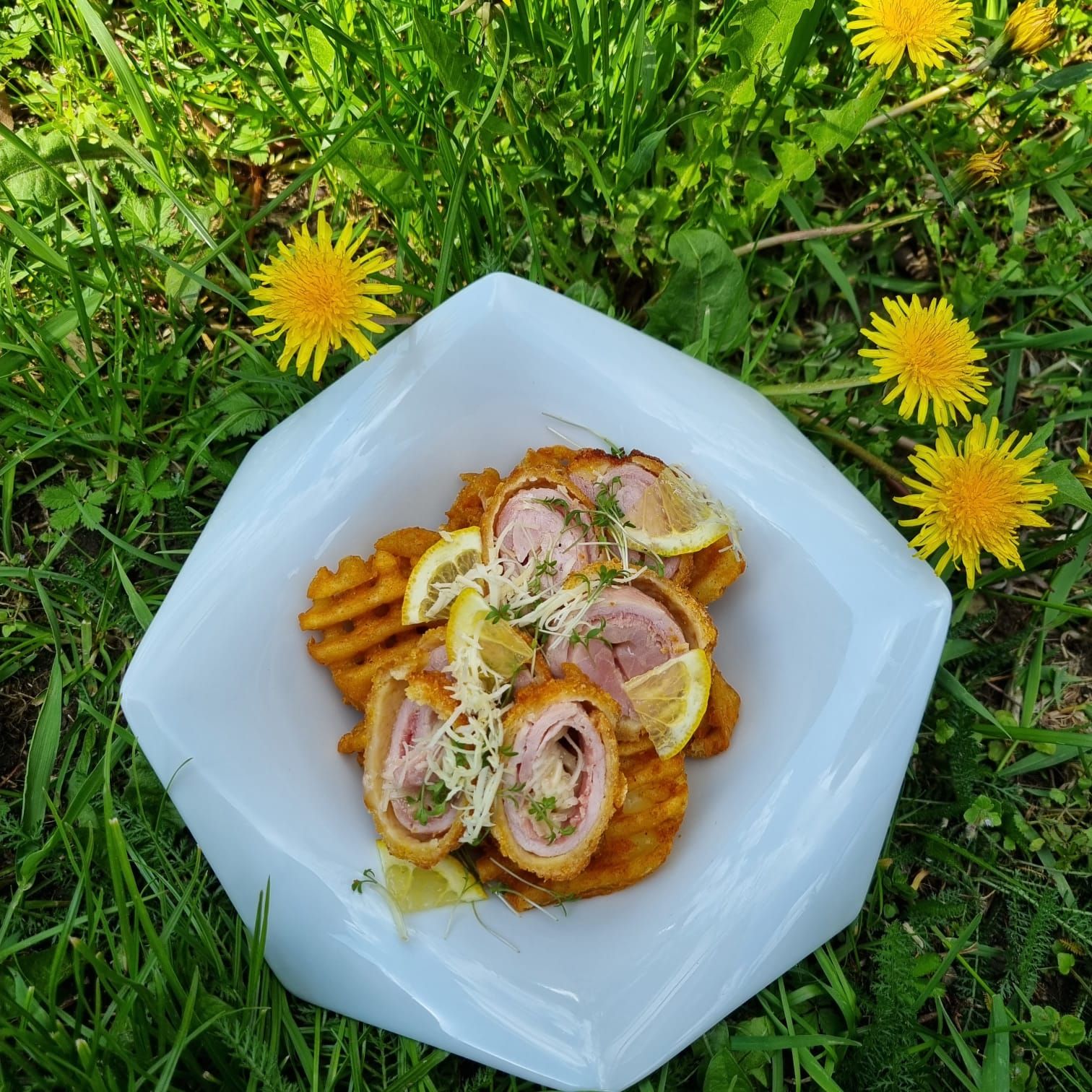 Egy tányér kordon tekercs és gofrisült krumpli, citromszeletekkel és zsázsával díszítve, egy pitypangos réten fekszik.