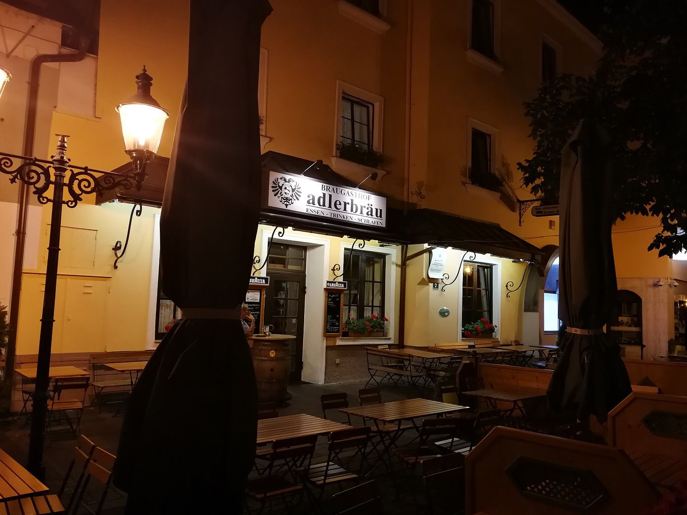 Éjszakai felvétel egy "Adlerbräu" feliratú étteremről, kint üres asztalokkal.