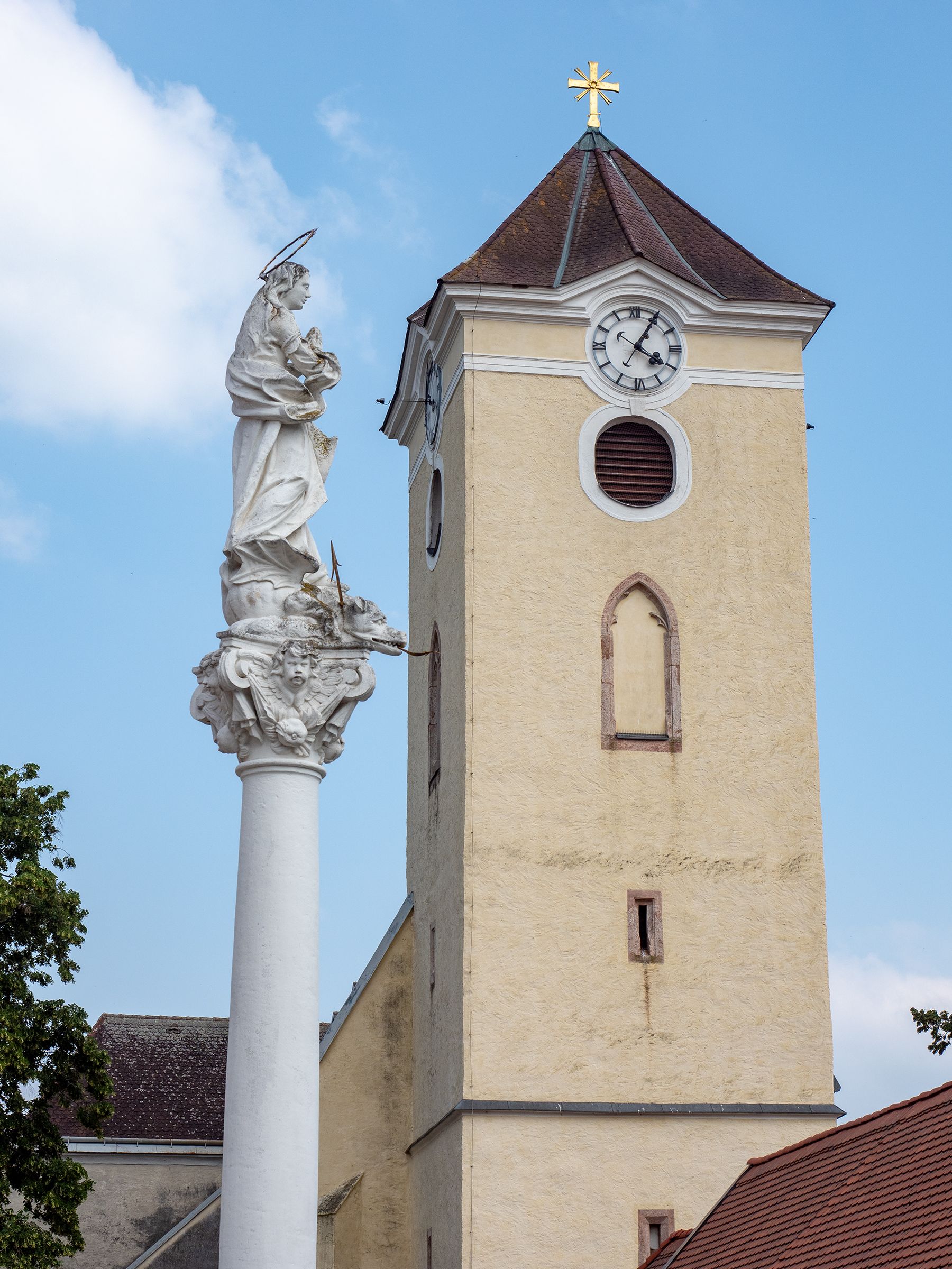 Templomtorony órával és Mária-oszloppal Schrattenthalban, kék égbolt előtt.