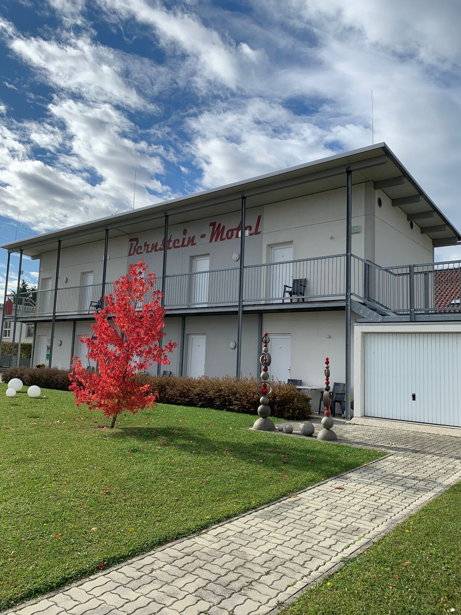 Kétemeletes épület "Bernstein Motel" felirattal, előtérben piros fa, kék égbolt felhőkkel.
