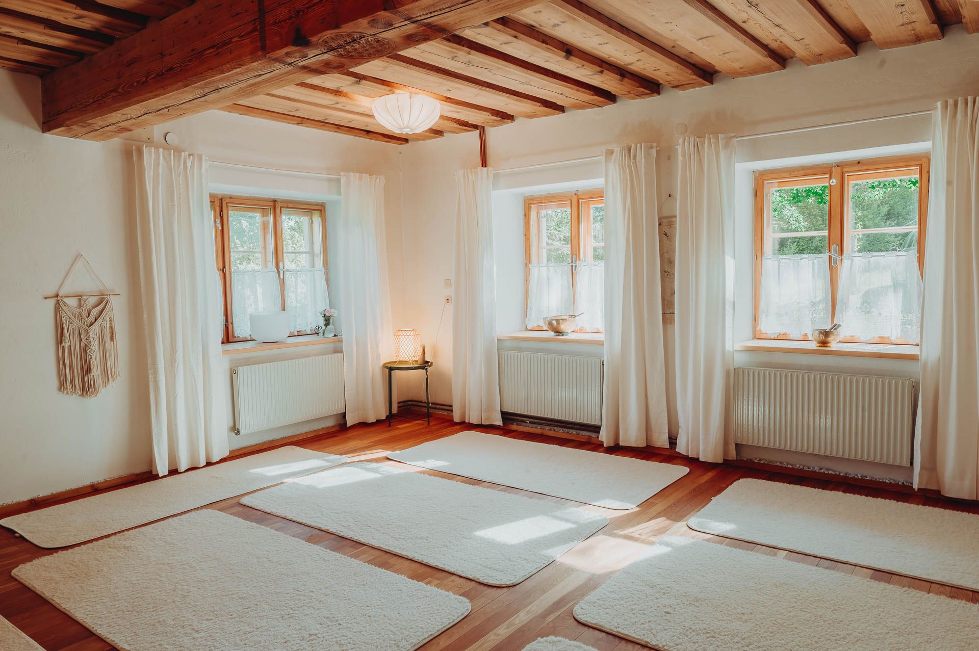 Világos jógaterem fapadlóval, fehér szőnyegekkel és nagy ablakokkal, függönyökkel.