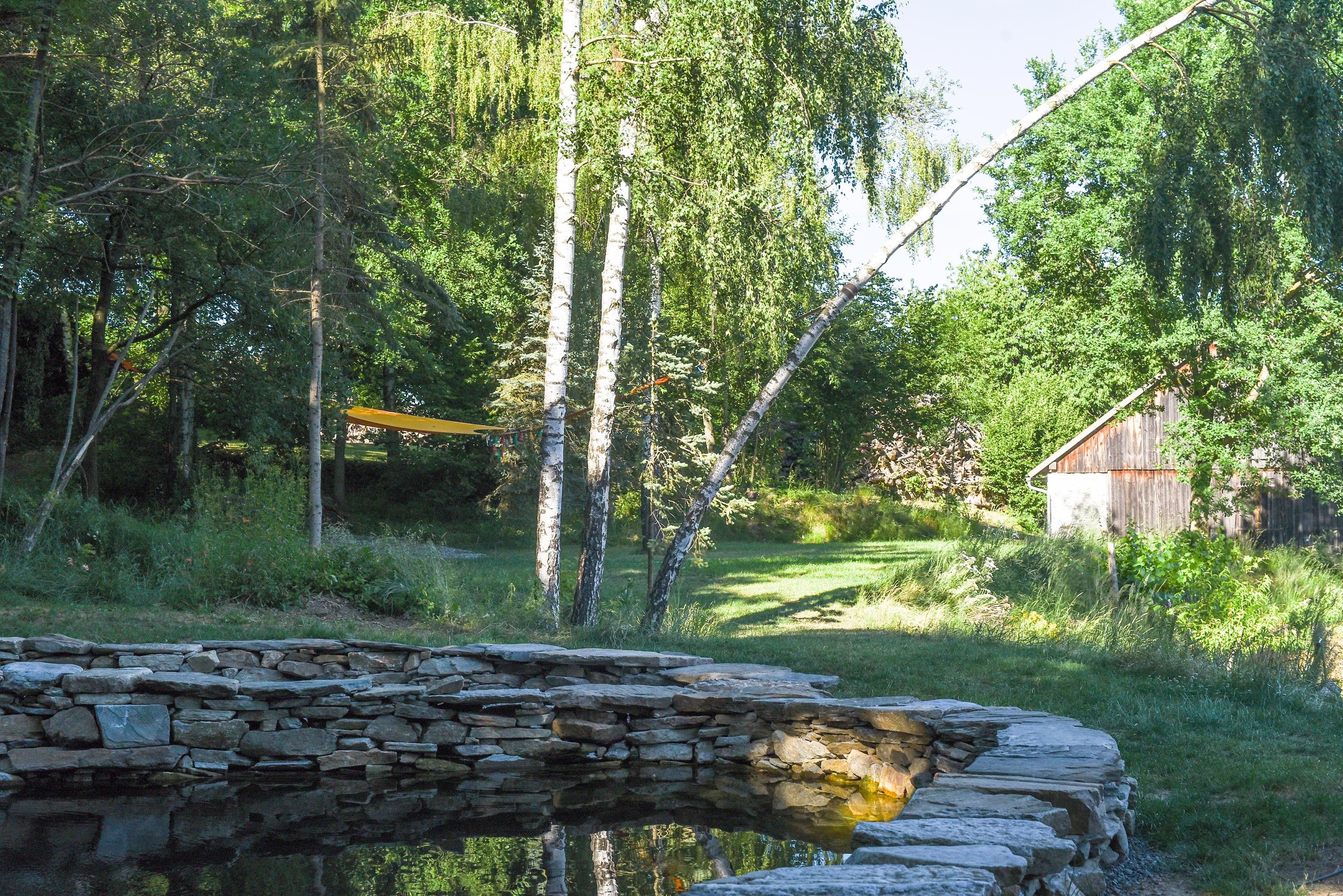 Egy kert, amelyben egy természetes kövekből kialakított kis tó található, fákkal körülvéve, a háttérben pedig egy faház látható.