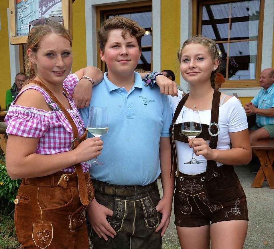 Három fiatalember hagyományos bajor ruhában, borospohárral egy sárga épület előtt.