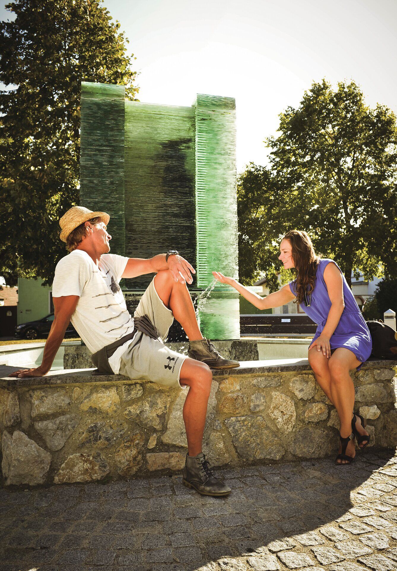 Egy pár ül a falon egy szökőkút előtt. A nő a vízzel játszik. 