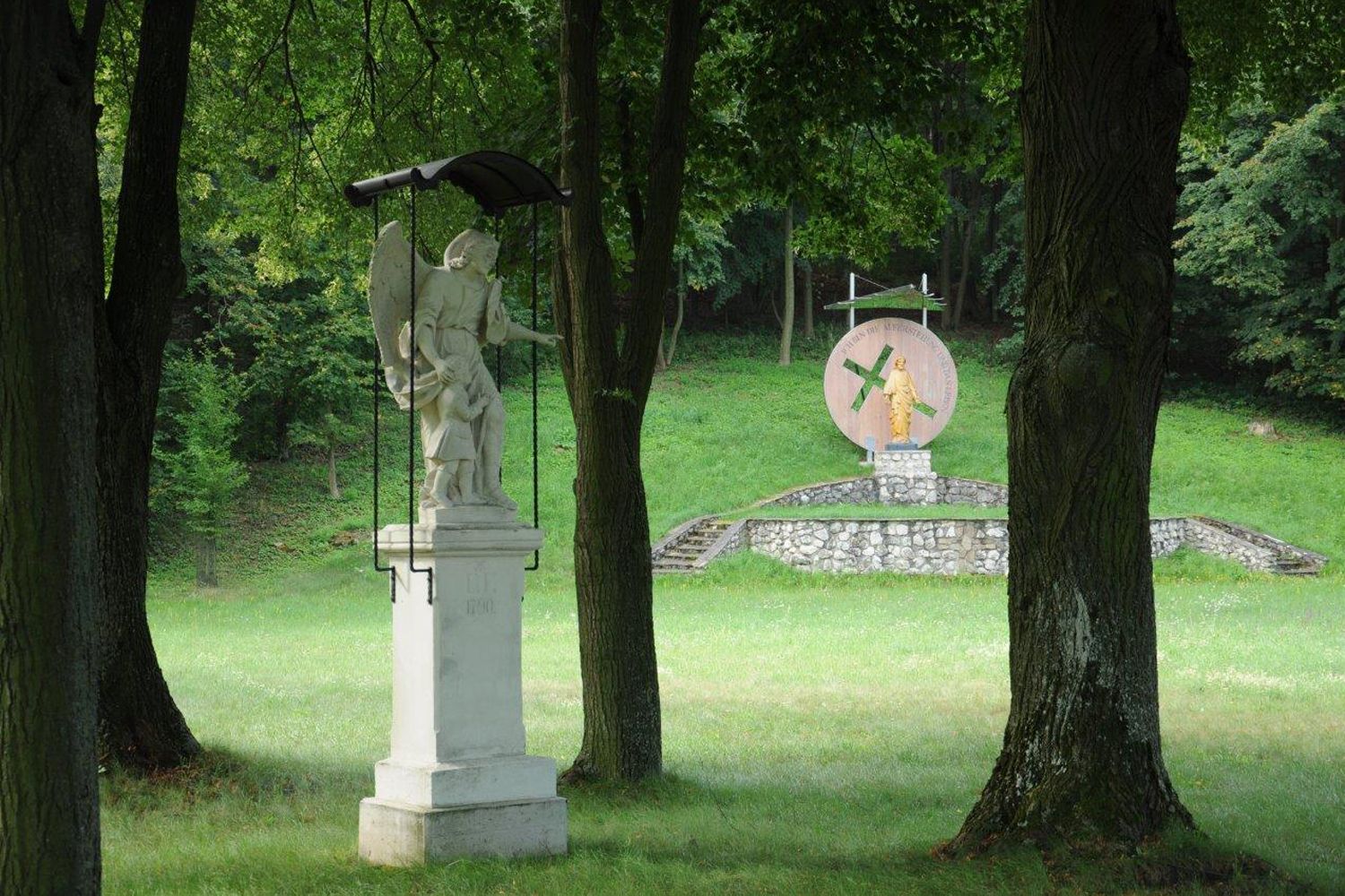 Angyalszobor gyermekével az előtérben, a háttérben egy másik szobor egy talapzaton egy erdős parkban.