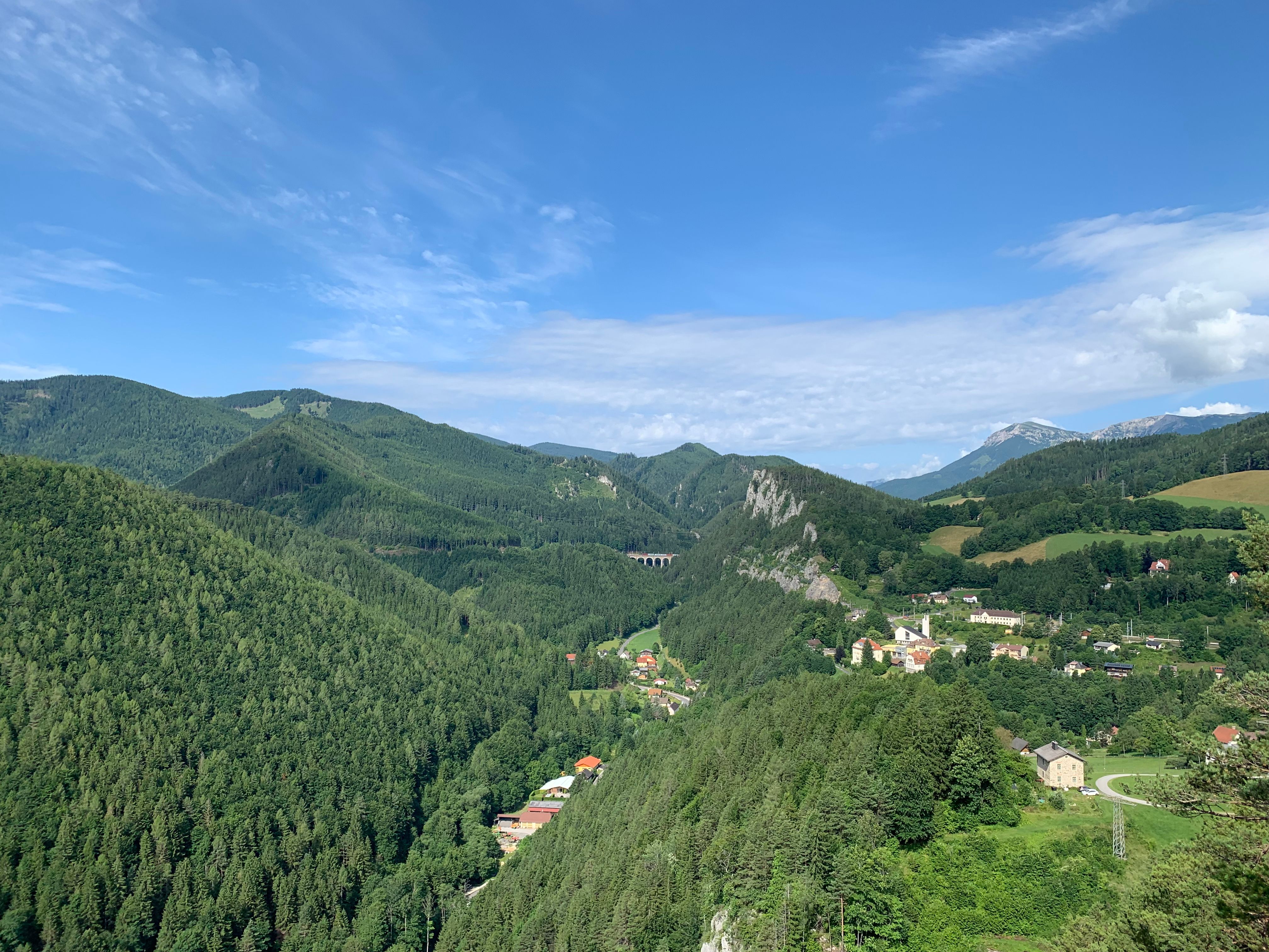 Panorámakép egy zöld hegyvidéki tájra egy kis faluval és egy viadukttal.