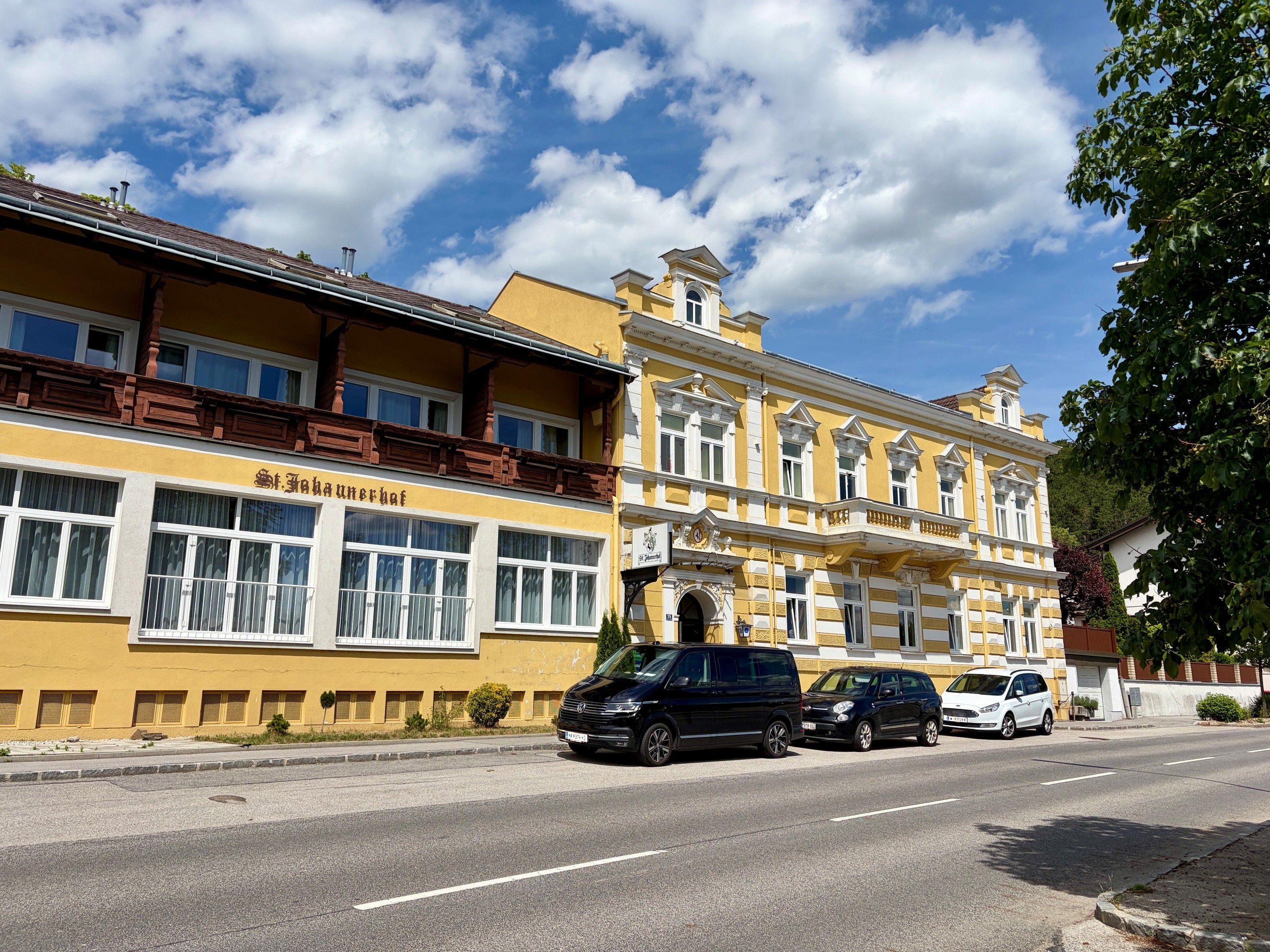 Sárga épület "St. Johannerhof" felirattal egy úton, körülötte parkoló autók és fák.