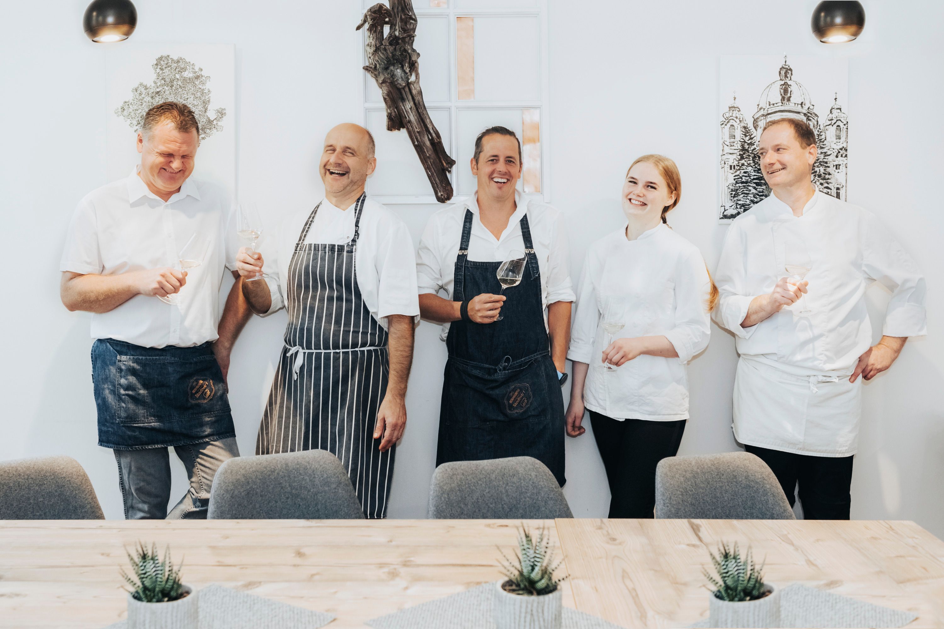 Öt séfnek öltözött ember nevetgél, kezükben borospoharakkal.