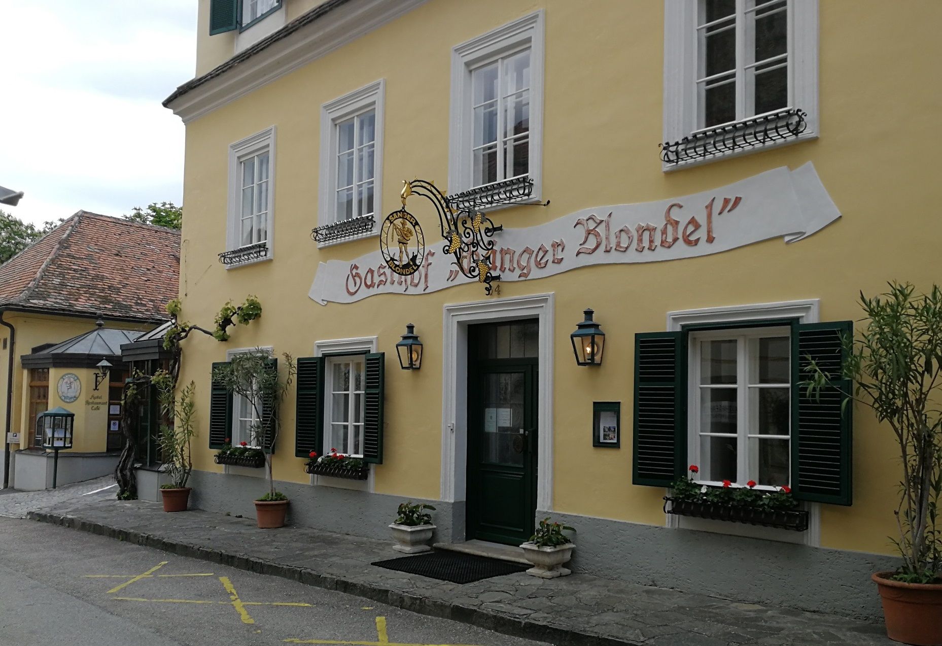 Sárga épület "Gasthof Sänger Blondel" felirattal, zöld redőnyös ablakokkal és virágládákkal.