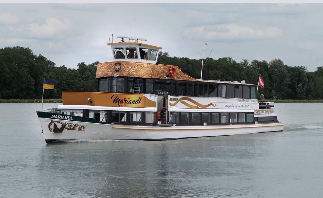 Az MS Mariandl nevű kirándulóhajó egy fákkal körülvett folyón hajózik. Ukrajna és Ausztria zászlaját viseli.