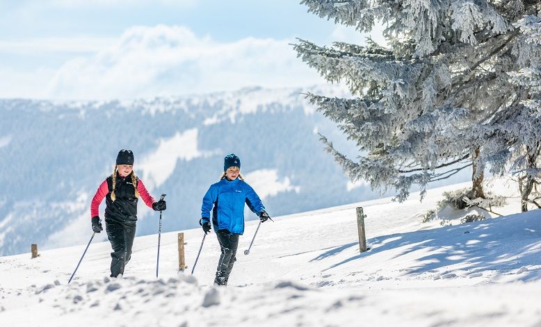 Két ember sífutás egy hóval borított pályán, a háttérben hegyek.