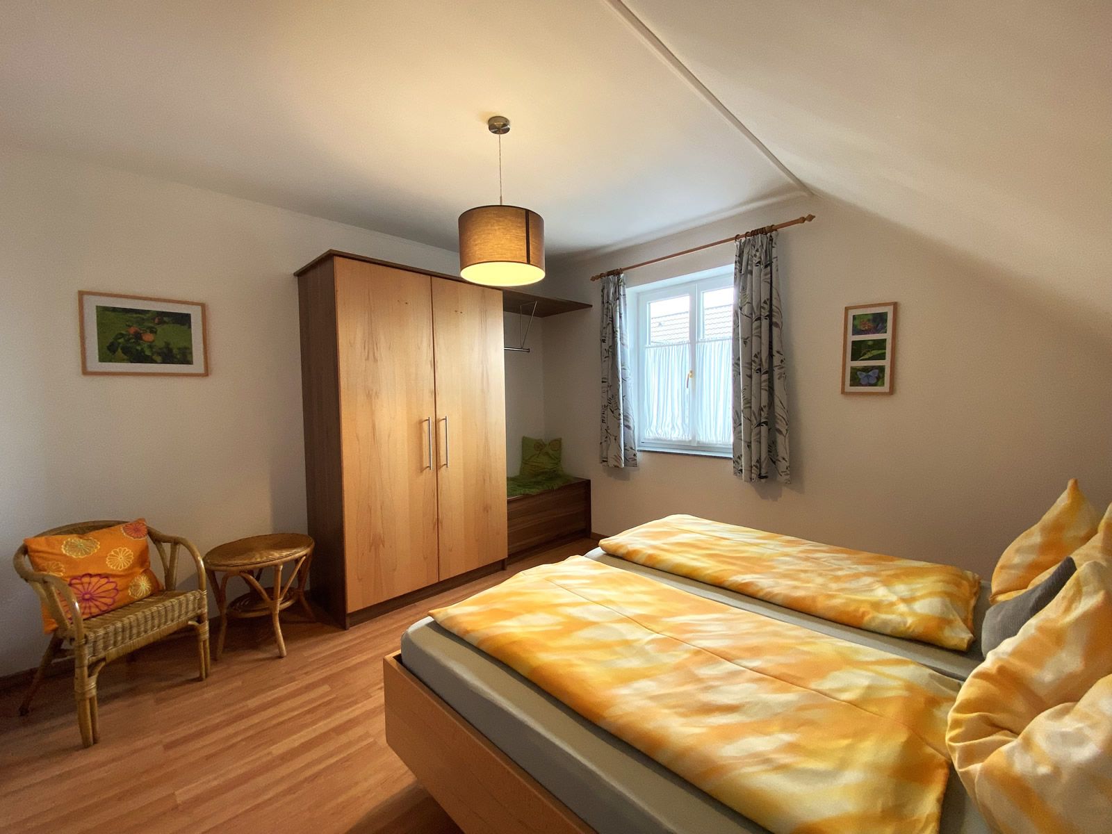 Hálószoba franciaággyal, fa szekrénnyel, fonott székkel és függönyös ablakkal.