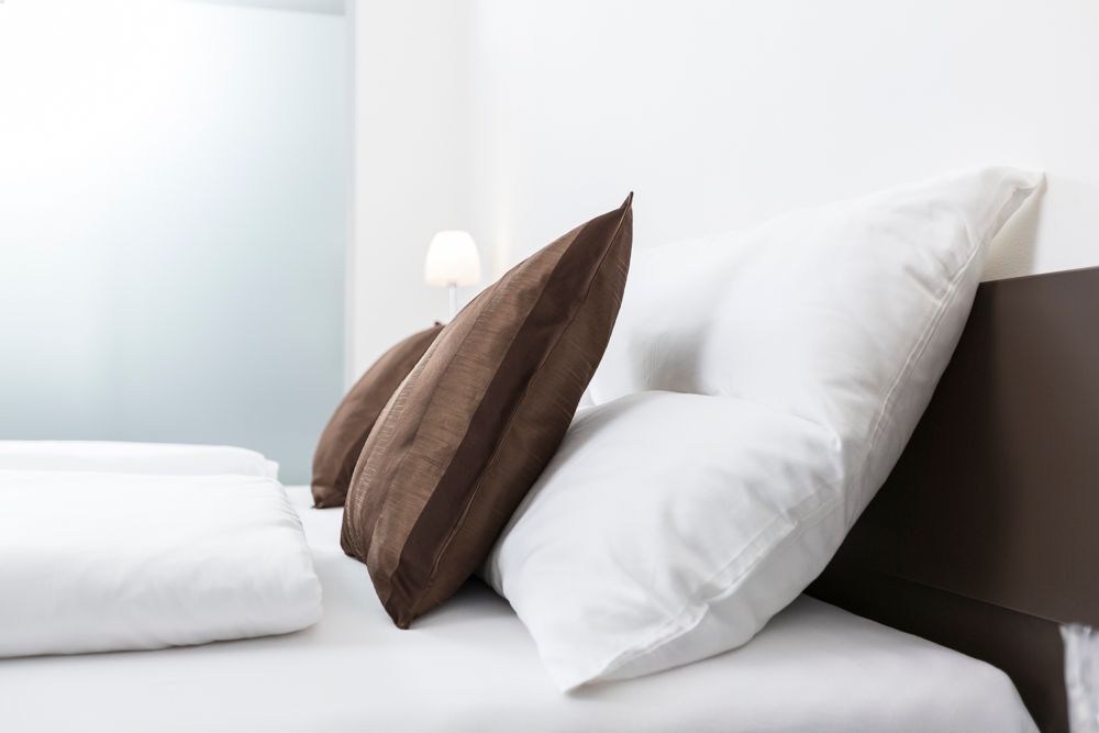 Egy ágy fehér párnákkal és barna díszpárnákkal, a háttérben egy lámpa.