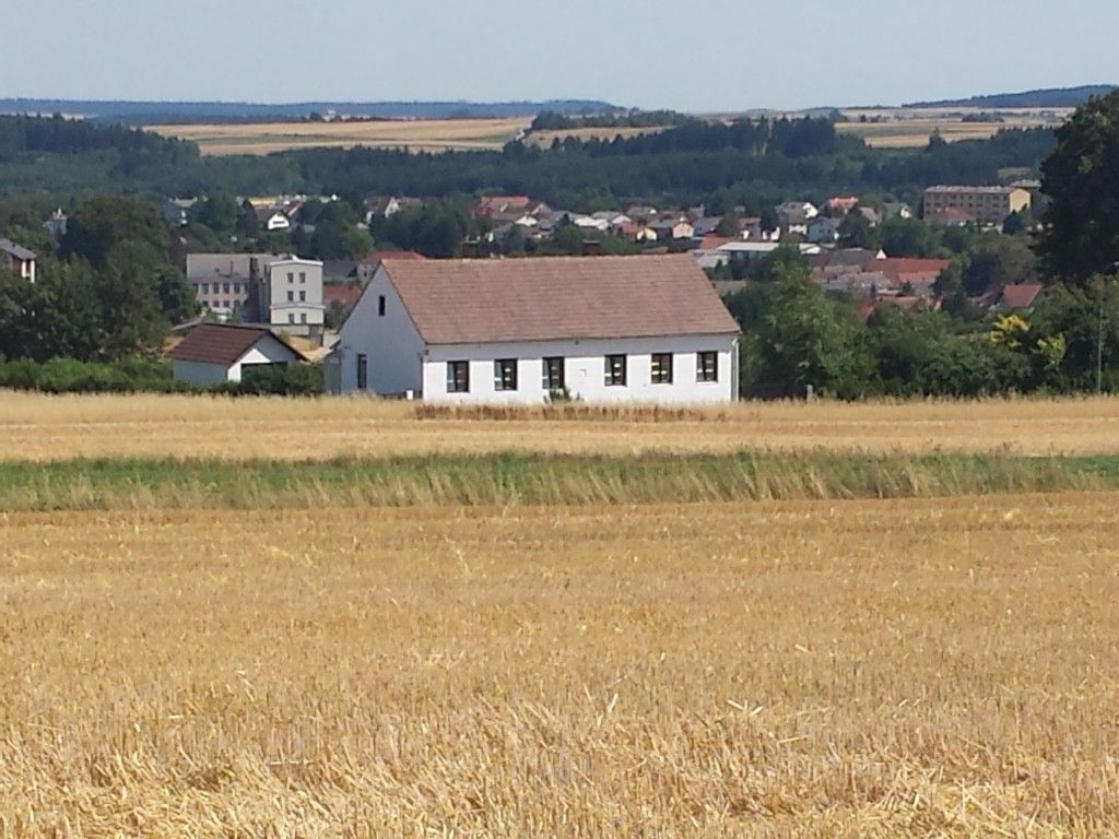Egy piros tetejű fehér ház áll egy mezőn, a háttérben egy város és egy erdő.