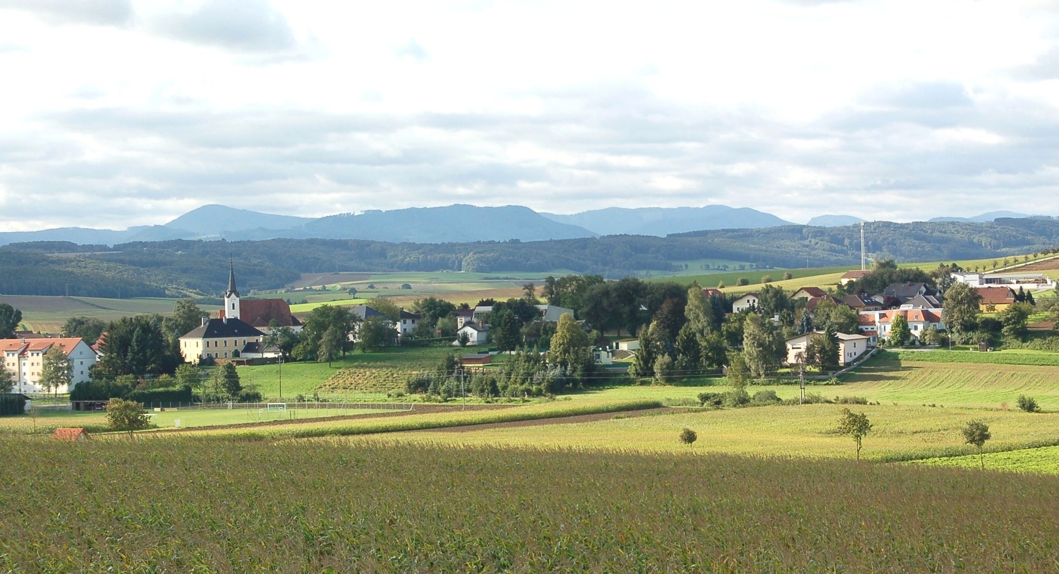Tájkép egy faluval és templommal St. Margarethen an der Sierningben, mezőkkel és dombokkal körülvéve.