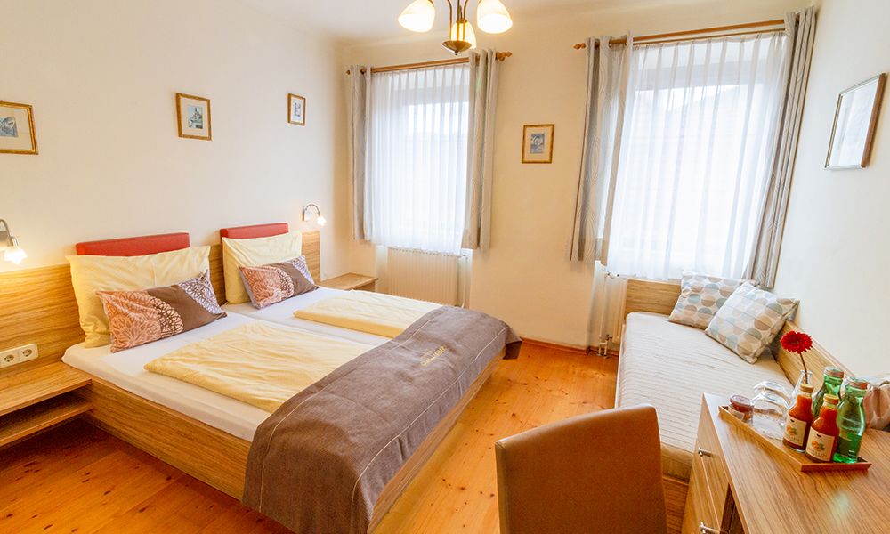 Hangulatos vendégszoba franciaággyal, napozóággyal, fabútorokkal és díszpárnákkal. Világos színű függönyök és képek a falakon.