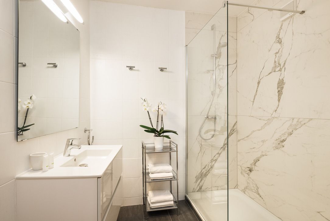 Modern fürdőszoba fehér mosdóval, tükörrel, üvegzuhannyal és orchideával a polcon.