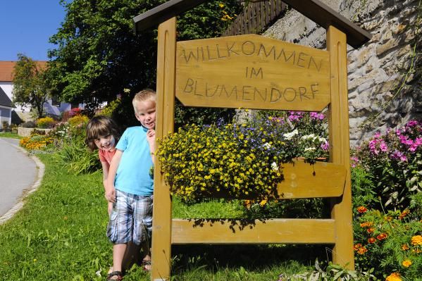 Két gyermek áll egy fából készült tábla mellett, amelyen az "Üdvözöljük a virágfaluban" felirat olvasható, körülötte színes virágok.